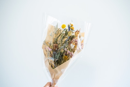 A bouquet of dried flowers wrapped in clear cellophane and brown paper. The arrangement includes various types and colors of flowers, such as yellow billy balls, white daisies, and pink statice. The bouquet is held up against a plain, light-colored background.