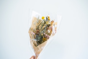 A bouquet of dried flowers wrapped in clear cellophane and brown paper. The arrangement includes various types and colors of flowers, such as yellow billy balls, white daisies, and pink statice. The bouquet is held up against a plain, light-colored background.