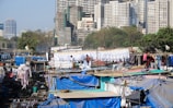 A densely packed urban area with numerous makeshift structures covered in tarps and clothing hanging out to dry. Several people are engaged in laundry activities. A backdrop of high-rise buildings looms over the scene, with scattered greenery among the buildings.