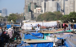 A densely packed urban area with numerous makeshift structures covered in tarps and clothing hanging out to dry. Several people are engaged in laundry activities. A backdrop of high-rise buildings looms over the scene, with scattered greenery among the buildings.
