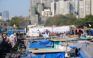 A densely packed urban area with numerous makeshift structures covered in tarps and clothing hanging out to dry. Several people are engaged in laundry activities. A backdrop of high-rise buildings looms over the scene, with scattered greenery among the buildings.