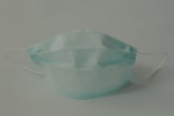 A single disposable surgical mask with pleats is positioned against a plain background. The mask features adjustable ear loops and a blue color.
