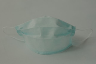 A single disposable surgical mask with pleats is positioned against a plain background. The mask features adjustable ear loops and a blue color.