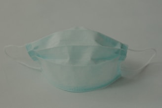 A single disposable surgical mask with pleats is positioned against a plain background. The mask features adjustable ear loops and a blue color.