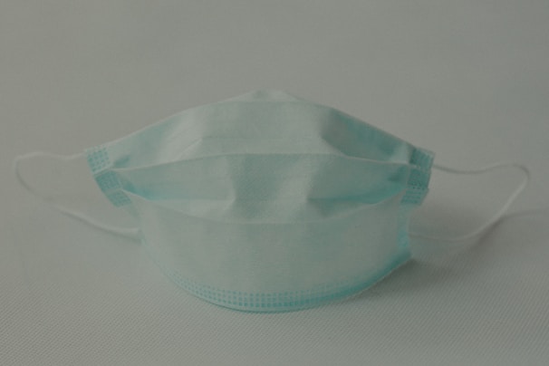 A single disposable surgical mask with pleats is positioned against a plain background. The mask features adjustable ear loops and a blue color.
