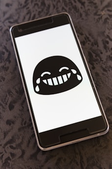 A smartphone displays a black and white laughing emoji with tears of joy on its screen. The phone lies on a textured dark surface, creating a contrast between the screen's brightness and the background.