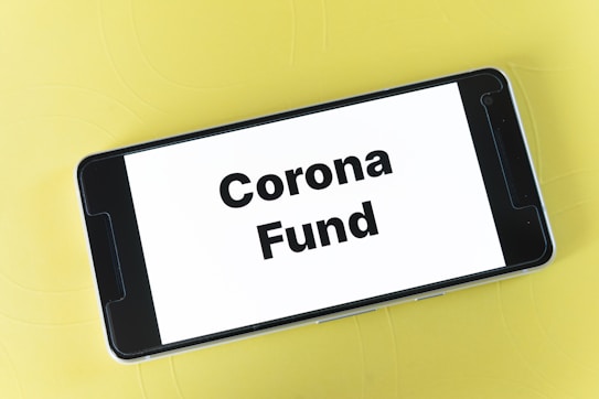 A smartphone displaying the text 'Corona Fund' on its screen is placed on a light yellow surface.