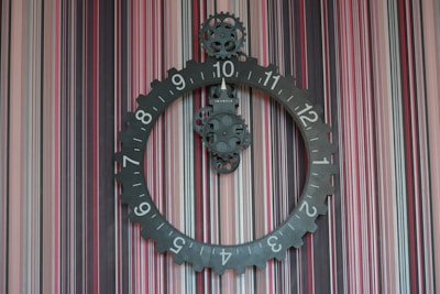 A large, circular, gear-shaped clock with visible mechanical components hangs against a background of multicolored vertical stripes. The numbers on the clock are arranged around the perimeter, and there are gear mechanisms visible within the center.