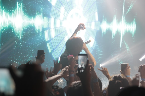 An energetic concert scene with a performer holding a microphone facing a cheering crowd. The background features vibrant digital visuals with radial patterns and green sound waves. Many audience members are holding up smartphones to capture the performance.