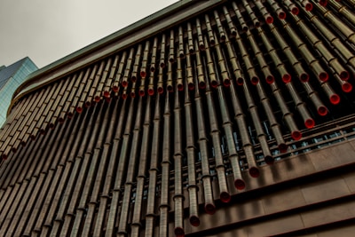 A close-up view of a modern building facade, featuring an array of long, cylindrical pipes with red inner linings. The pipes are neatly aligned in a vertical pattern, creating a striking architectural feature. The building's structure has a metallic appearance with a contrasting glass segment visible on the left.
