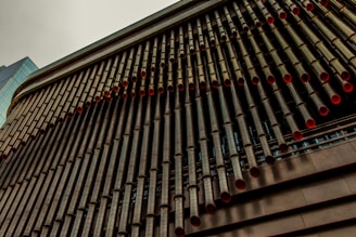 A close-up view of a modern building facade, featuring an array of long, cylindrical pipes with red inner linings. The pipes are neatly aligned in a vertical pattern, creating a striking architectural feature. The building's structure has a metallic appearance with a contrasting glass segment visible on the left.