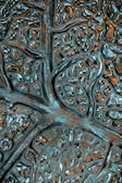 A close-up of an abstract textured surface featuring irregular patterns resembling neural networks or biological structures. The surface appears metallic with reflective qualities and intricate grooves.