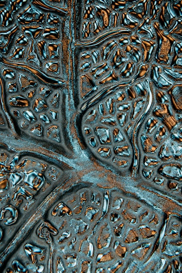 A close-up of an abstract textured surface featuring irregular patterns resembling neural networks or biological structures. The surface appears metallic with reflective qualities and intricate grooves.