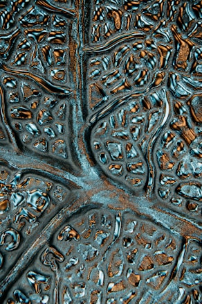A close-up of an abstract textured surface featuring irregular patterns resembling neural networks or biological structures. The surface appears metallic with reflective qualities and intricate grooves.