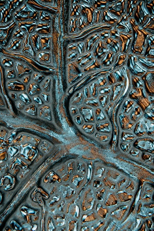 A close-up of an abstract textured surface featuring irregular patterns resembling neural networks or biological structures. The surface appears metallic with reflective qualities and intricate grooves.