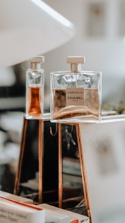 A close-up of two Chanel perfume bottles displayed on a reflective surface. The larger bottle prominently features the label 'Gabrielle Chanel Paris' and contains a light amber liquid. The background is softly blurred, creating an elegant and luxurious atmosphere.