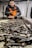 A person wearing an orange safety vest and black gloves is sorting and handling a large number of razor clams on a metal processing table in an industrial setting.