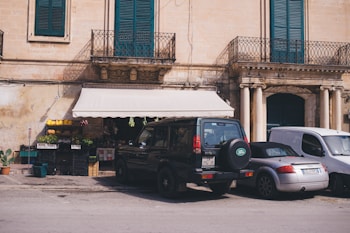 A small outdoor market scene with a display of fruits and vegetables under a white awning attached to an old building. There are parked cars in front, including a dark green Land Rover and a silver Audi. The building facade features ornate columns and blue shutters.