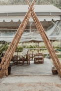 A rustic outdoor seating area with wicker chairs and tables arranged on a concrete surface, partially enclosed by a tent-like structure made of bamboo poles. The area is surrounded by greenery, contributing to a peaceful and natural atmosphere.