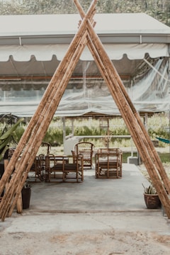 A rustic outdoor seating area with wicker chairs and tables arranged on a concrete surface, partially enclosed by a tent-like structure made of bamboo poles. The area is surrounded by greenery, contributing to a peaceful and natural atmosphere.