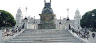 A grand stone monument with a statue is positioned at the top of wide marble steps. The background features an ornate, historical building with domes and elaborate architecture. Two large trees and several lampposts line the sides of the structure, adding to the classical ambiance.