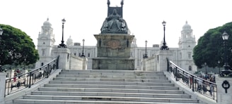 A grand stone monument with a statue is positioned at the top of wide marble steps. The background features an ornate, historical building with domes and elaborate architecture. Two large trees and several lampposts line the sides of the structure, adding to the classical ambiance.