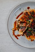 Crispy dumplings topped with chopped green onions and drizzled with a dark, savory sauce are arranged on a round, grayish plate. The dumplings have a golden-brown texture and are sliced open, revealing the filling inside. The arrangement is artistic, creating an appetizing visual appeal.