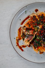 Crispy dumplings topped with chopped green onions and drizzled with a dark, savory sauce are arranged on a round, grayish plate. The dumplings have a golden-brown texture and are sliced open, revealing the filling inside. The arrangement is artistic, creating an appetizing visual appeal.