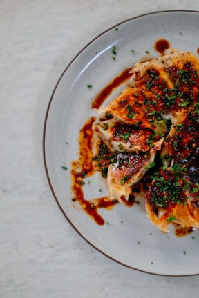 Crispy dumplings topped with chopped green onions and drizzled with a dark, savory sauce are arranged on a round, grayish plate. The dumplings have a golden-brown texture and are sliced open, revealing the filling inside. The arrangement is artistic, creating an appetizing visual appeal.