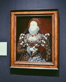 A detailed portrait of a historical figure, dressed in an ornate and elaborately decorated gown with intricate patterns and lace. The attire includes a ruffled collar and voluminous sleeves, with a background of a dark maroon hue. The portrait is housed in a wooden frame and accompanied by a small informational plaque on the wall.