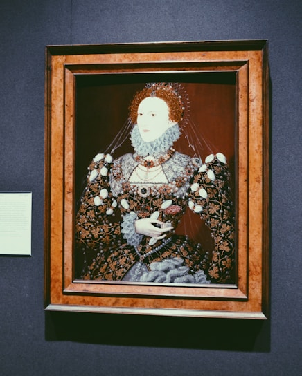 A detailed portrait of a historical figure, dressed in an ornate and elaborately decorated gown with intricate patterns and lace. The attire includes a ruffled collar and voluminous sleeves, with a background of a dark maroon hue. The portrait is housed in a wooden frame and accompanied by a small informational plaque on the wall.