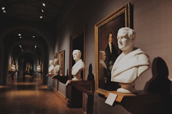 A dimly lit museum hallway features classical sculptures and framed portraits along the walls. The busts are elegantly mounted on wooden shelves, and there is a sense of depth with the corridor extending into the distance. The artworks are meticulously arranged, creating a symmetrical and balanced visual experience.