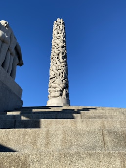 A tall, intricately carved stone monolith features numerous intertwined human figures. The structure stands on a platform of stone steps, with a clear blue sky in the background. Part of another stone sculpture is visible to the left.