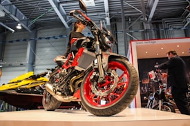 A sleek, modern motorcycle with striking red and black details is prominently displayed on a platform indoors. Nearby, people are interacting with the motorbike, suggesting an exhibition or trade show setting. Overhead, the building&rsquo;s industrial ceiling and lighting fixtures are visible.