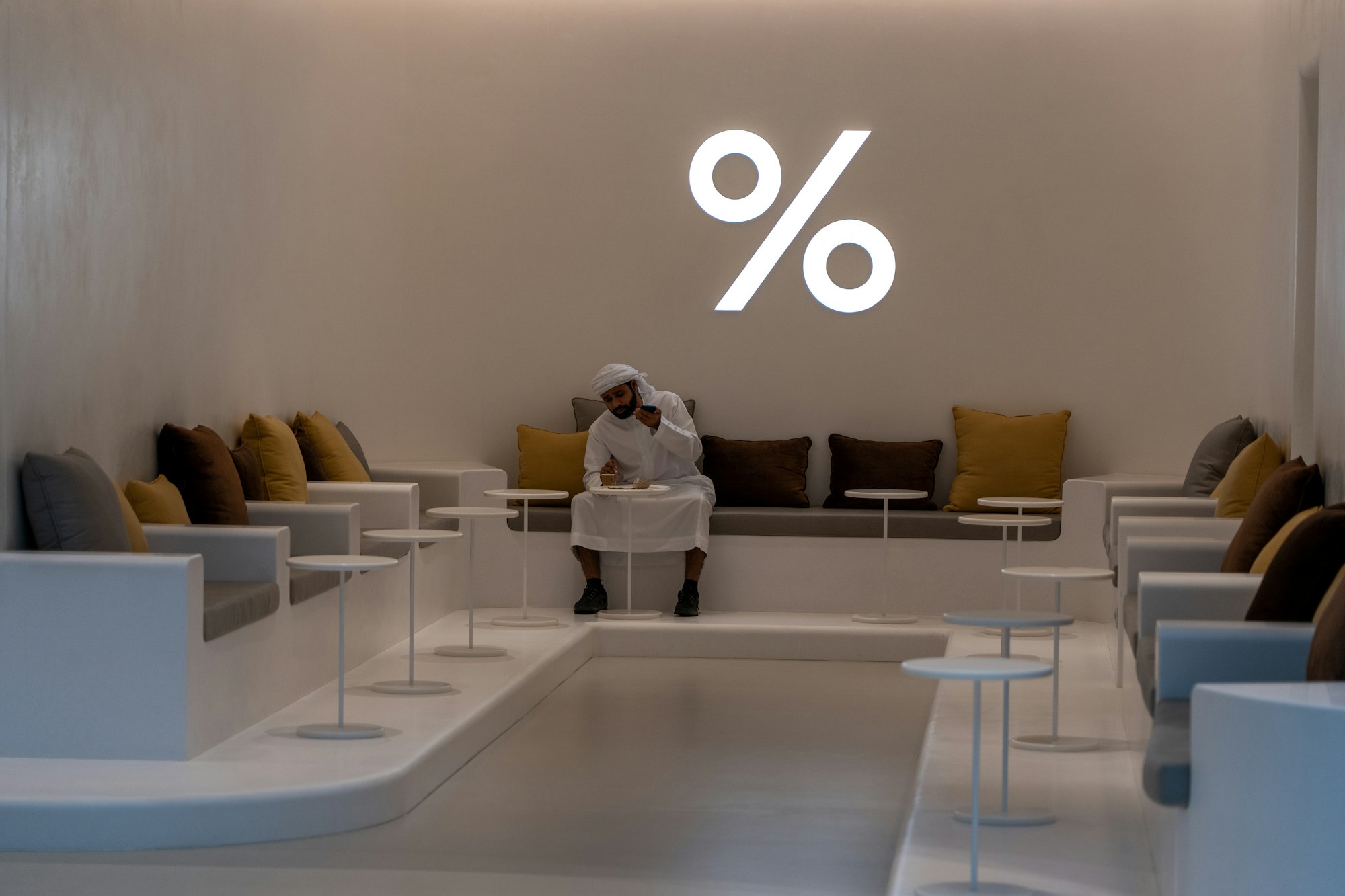 A man having a coffee in one of the coffee shops in the Dubai Mall, UAE.  I loved the white of his traditional outfit, against the white of the floor, tables and percentage symbol.