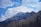 A rugged, snow-capped mountain peak rises sharply against a vibrant blue sky filled with fluffy white clouds. The jagged edges of the mountain are highlighted by patches of snow and shadow, creating a striking contrast with the sky.