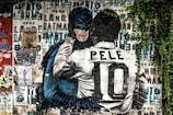 A vibrant mural features Batman embracing a soccer player with the name Pelé and the number 10 on his back. The background is a collage of various posters and graffiti with a mix of text and images. Green foliage partially covers the mural on the right side, adding a touch of nature to the urban art scene.