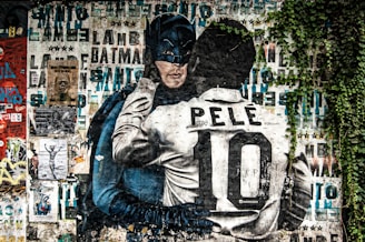 A vibrant mural features Batman embracing a soccer player with the name Pelé and the number 10 on his back. The background is a collage of various posters and graffiti with a mix of text and images. Green foliage partially covers the mural on the right side, adding a touch of nature to the urban art scene.