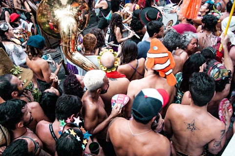A vibrant crowd of people closely gathered in a festive atmosphere. Many individuals are shirtless, and some wear colorful accessories. A large brass tuba can be seen in the scene, adding to the lively energy. Costume elements like a clownfish hat and paper stars are visible, suggesting a carnival or parade setting.