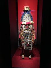 The image displays a richly ornate costume on display, prominently featuring intricate beadwork and vibrant colors. The costume is adorned with detailed patterns and symbols, set against a red background within a glass case.
