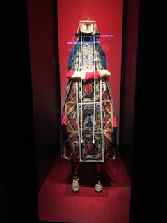 The image displays a richly ornate costume on display, prominently featuring intricate beadwork and vibrant colors. The costume is adorned with detailed patterns and symbols, set against a red background within a glass case.