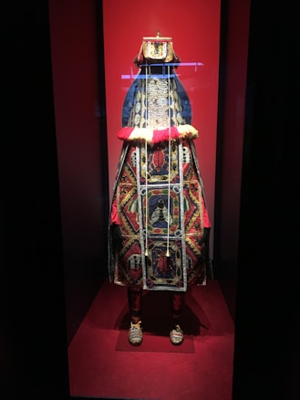 The image displays a richly ornate costume on display, prominently featuring intricate beadwork and vibrant colors. The costume is adorned with detailed patterns and symbols, set against a red background within a glass case.