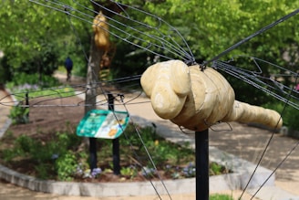 A wooden sculpture resembling an insect, possibly a bee or dragonfly, with metal wire wings is situated in an outdoor garden area. The background includes a pathway, vegetation, and signage. A person is walking in the distance, contributing to the natural setting of the scene.