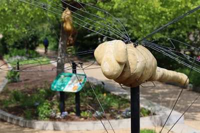 A wooden sculpture resembling an insect, possibly a bee or dragonfly, with metal wire wings is situated in an outdoor garden area. The background includes a pathway, vegetation, and signage. A person is walking in the distance, contributing to the natural setting of the scene.