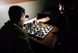 Two people are engaged in a chess game, sitting opposite each other in a dimly lit room. The focus is on the chessboard, which has both black and white pieces arranged on it. One player is making a move while holding a pen in their other hand, and the other player is observing the move attentively. The lighting creates shadows, giving the scene an intense and focused atmosphere.