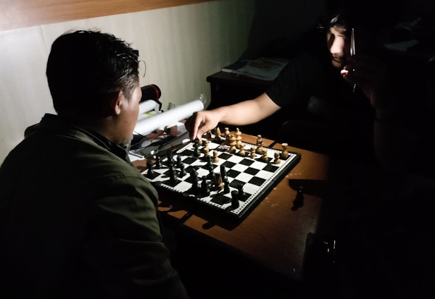 Two people are engaged in a chess game, sitting opposite each other in a dimly lit room. The focus is on the chessboard, which has both black and white pieces arranged on it. One player is making a move while holding a pen in their other hand, and the other player is observing the move attentively. The lighting creates shadows, giving the scene an intense and focused atmosphere.