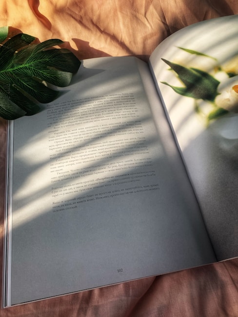 A book with text written in a language using the Cyrillic script is open, slightly lying on a soft, light pink fabric. Sunlight filters through what appears to be a leaf, casting shadows and creating a calming pattern on the pages and fabric.