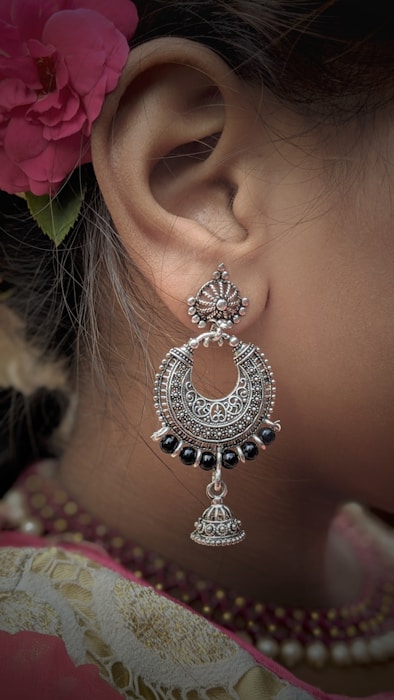 An intricately designed silver earring with black beads complements a traditional attire. A red floral hair accessory adds a touch of elegance, with fine detailing visible on both the earring and the fabric.