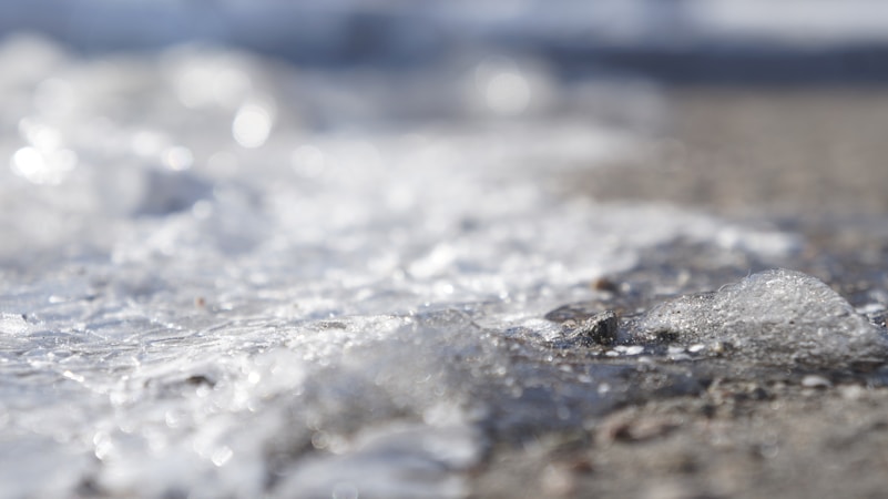 A close-up of melting ice particles on a flat surface, with a thin layer of water creating a blurry, reflective quality. The ice appears textured and interspersed with small rocks and dirt, with sunlight reflecting off its surface.