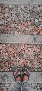 A pair of feet wearing black sandals is positioned on a gravel path with rough, uneven stones. The stones have a mix of grey and reddish-brown hues and are bordered by narrow concrete strips.